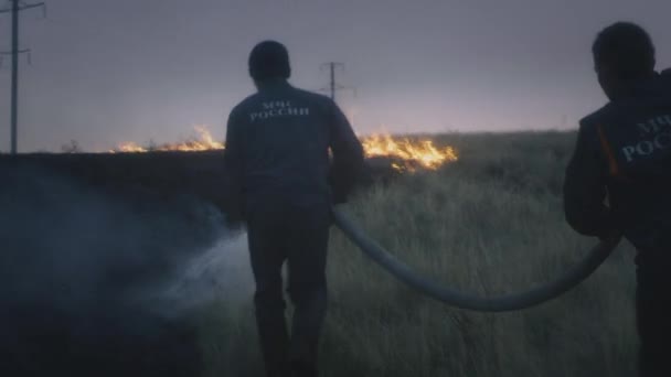 Dos bomberos reales llevan una manguera con agua en las manos, apagan la llama ardiente. La naturaleza está en llamas: hierba, estepa y campo — Vídeos de Stock