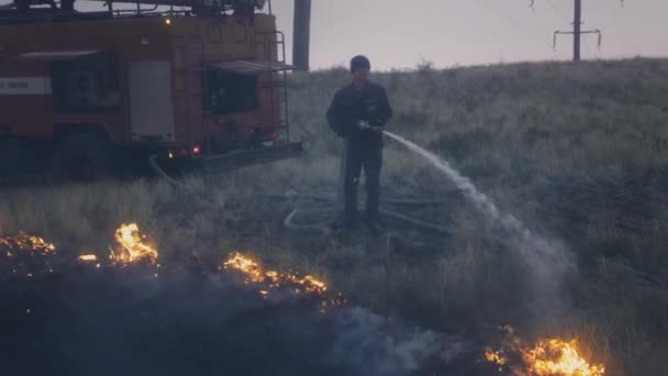 UFA RUSSIA - 07.08.2021: Um verdadeiro bombeiro apaga uma chama ardente de uma mangueira com água. A natureza está a arder. A água de uma mangueira é pulverizada no fogo. Há um carro de bombeiros por perto.. — Vídeo de Stock