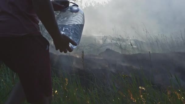 A volunteer man or boy extinguishes a wild fire. Pours water from a plastic bottle. Rain forest wildfire disaster, dry bushes burning, fire reasons. — Stock Video