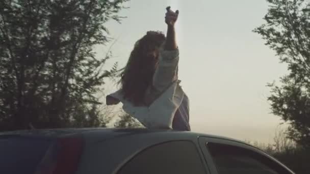 A fiatal, gyönyörű lány felemelt kézzel ül az autóban, egy nő a kocsi napfénytetőjén. Élvezi az életet naplementekor egy autó nyílása — Stock videók
