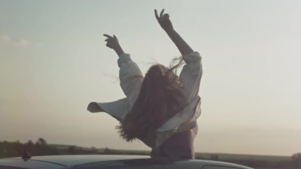 Joven hermosa chica monta en el coche con las manos levantadas hacia arriba, una mujer en el techo corredizo del coche. Disfrutando de la vida al atardecer en una escotilla de coche — Vídeos de Stock