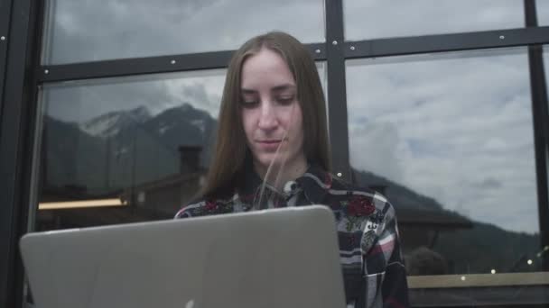 En ung frilansarflicka jobbar på en laptop på ett café nära bergen. Kvinnan skriver på en bärbar dator, program. — Stockvideo