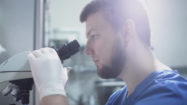 Un om de știință doctor într-o haină albastră se uită prin ochelarii unui microscop cu ochii. Setează lentile pentru fotografia macro — Videoclip de stoc