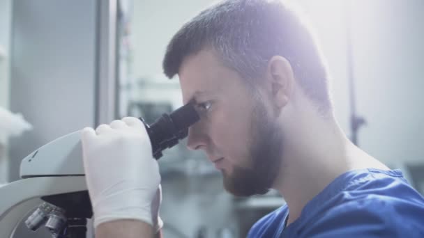 Een man dokter wetenschapper in een witte jas kijkt door de ogen van een microscoop met zijn ogen. Stelt lenzen op voor macrofotografie van cellen — Stockvideo