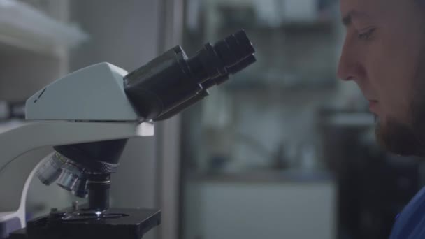 파란색 외투를 입은 남자 의사 과학자는 눈으로 현미경의 눈을 들여다보고 있다. 매크로 사진을 찍을 렌즈를 설치 한다 — 비디오