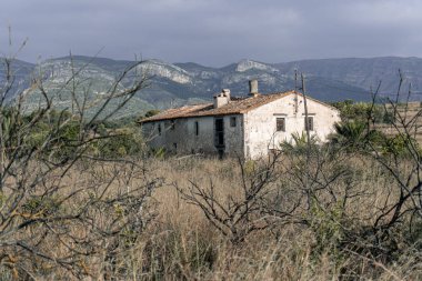 Tarlada terk edilmiş eski bir çiftlik evi, arkasında dağlar var..