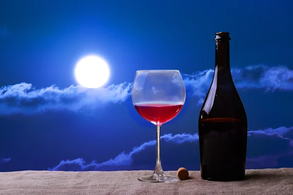 Бутылка и бокал красного вина на льняном столе против красочной луны заката — стоковое фото