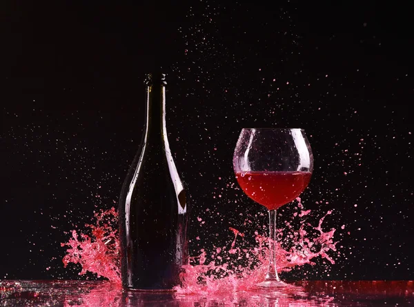 Bottle and glass with red wine, red wine splash, wine pouring on table on dark black background, big splash around Glass and bottle of red wine splash on black