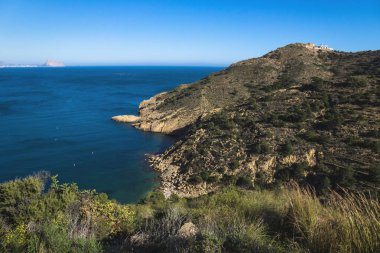 Doğal parktaki güneşli körfezde deniz feneri ile Serra Gelada ve arka planda Calpe 'nin Rock' Ifach 'ı, Albir, Costa Blanca, İspanya
