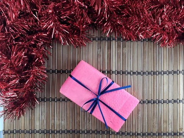 Kırmızı Tinsel ve Mavi Kurdeleli Pembe Hediye Kutusu Düz Yerde, Ahşap Arkaplan. Noel Süslemeleri ve Bambu Arkaplanlı Şimdiki Zaman 'ı Kopyala. Mutlu Noeller ve mutlu yıllar.