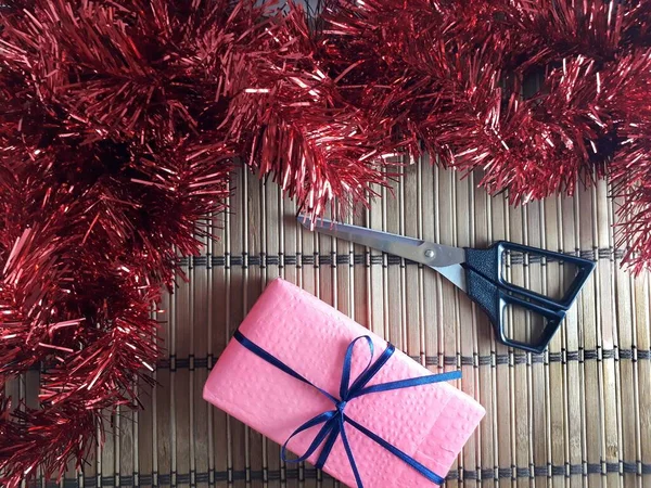 Mavi İnce Kurdeleli Pembe Hediye Kutusu. Tahta bambu üzerinde makaslı bir Noel hediyesi ve Red Tinsel. Yılın en güzel zamanında vermenin neşesi..