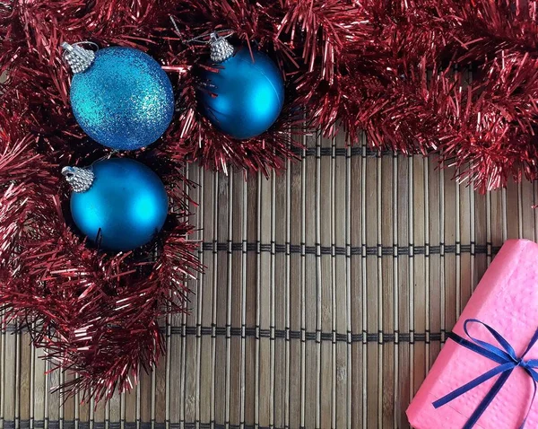 Kırmızı Tinsel, Mavi Baubles ve Mavi Kurdele ile sarılı Pembe Hediye Kutusu 'nun Düz Yatağı Görünümü. Noel Süslemeleri ve Bambu Arkaplanlı Şimdiki Zaman 'ı Kopyala. Mutlu Noeller ve mutlu yıllar.