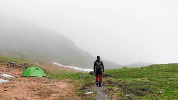 在一个雾蒙蒙的日子里 一个徒步旅行者独自在一座绿色帐篷附近的山上行走 — 图库视频影像