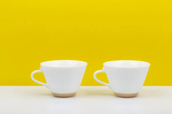 Dos tazas de cerámica brillante blanco sobre mesa blanca sobre fondo amarillo brillante —  Fotos de Stock