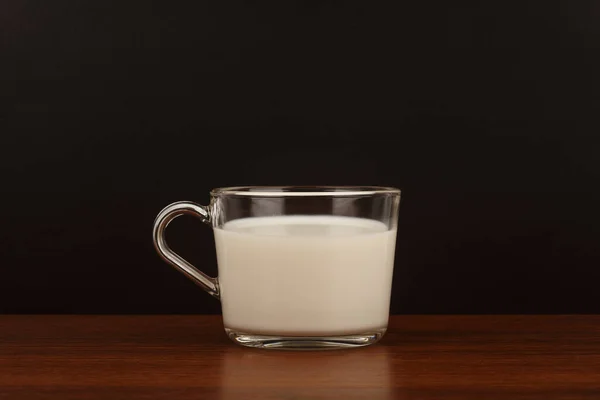 Transparante kop melk op bruine houten tafel tegen zwarte achtergrond met kopieerruimte — Stockfoto