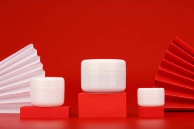 Kırmızı podyumlara konmuş krema kavanozları, dalgalarla süslenmiş kırmızı arka plana karşı. Lüks cilt bakımı kavramı