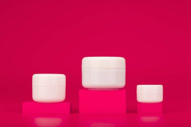 White unbranded cream jars on pink podiums against pink background with copy space. Face, neck and under eye products