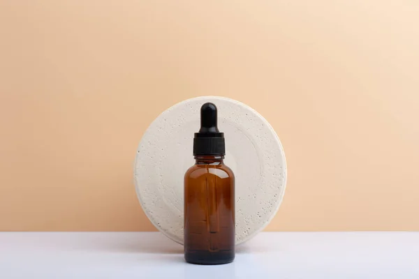 Skin serum in dark brown bottle against round stone circle on white table against beige background with copy space — Stock Photo, Image