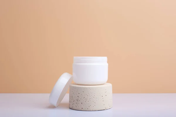 White opened cosmetic jar on beige podium and white table against beige background with copy space