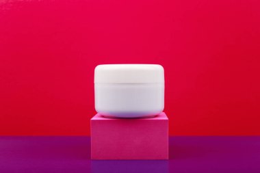 Kırmızı ve mor arkaplana karşı pembe kaide üzerine beyaz parlak kozmetik kavanozu