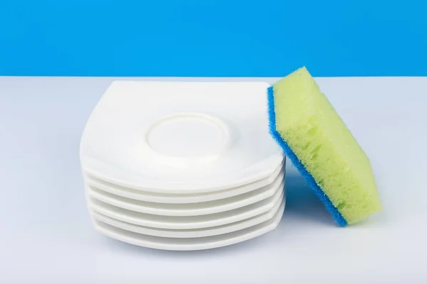Concept lave-vaisselle, vue grand angle de l'éponge de nettoyage et pile de soucoupes propres sur table blanche avec fond bleu — Photo