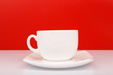 Beyaz seramik kahveyi ya da çay fincanını kırmızı arka planda beyaz masanın üzerinde çay tabağıyla kapat. Sıcak içecekler ve mutfak eşyaları kavramı