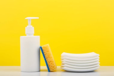 Renkli bulaşık konsepti, beyaz plastik şişede sıvı deterjan, sarı temizlik süngeri ve sarı arka plana karşı temiz tabaklar.