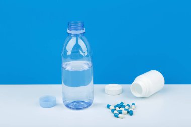 Yüksek açılı, mavi arka planda beyaz masaya dökülen haplarla birlikte su ve ilaç şişesi olan şeffaf plastik şişe. Tıbbi depresyon ve bipolar tedavi kavramı