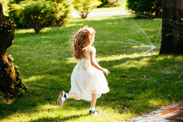Happy little girl running through the spray of water in the park — Stock Photo, Image