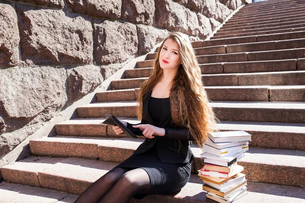 Beautiful girl student is sitting on stairs with tablet and stac — Stock Photo, Image