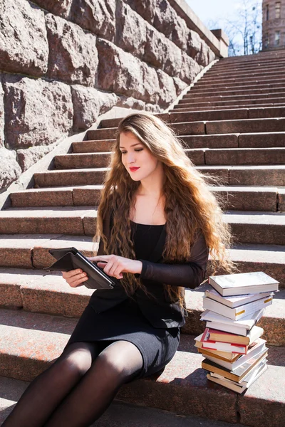 Beautiful girl student is sitting on stairs with tablet and stac — Stock Photo, Image