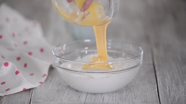 A woman adding egg yolks to whipped egg whites a mixing it with a spatula in a glass bowl. — Stock Video