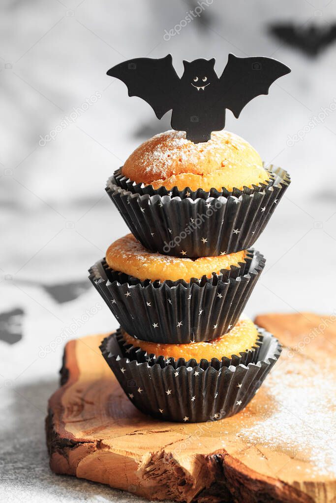 Halloween pumpkin muffins in black capsules decorated with cardboard bats. Festive Halloween cupcakes. Close-up view of delicious spooky halloween muffins on the grey background. Halloween recipes