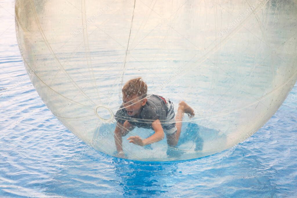 Child have fun inside big plastic balloon on the water of swimming pool on the summer resort. Little boy inside big inflatable transparent ball running and having fun. 