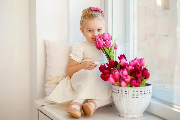 Vacker liten flicka i en vit klänning som sitter på en fönsterbräda w — Stockfoto