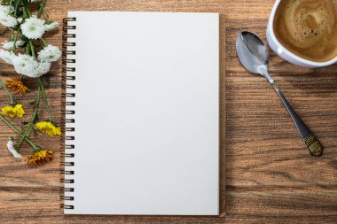 Tahta bir masanın üzerinde açık bir defter var. Kenarlarında çiçekler, yanında bir fincan kahve ve yanında bir çay kaşığı.