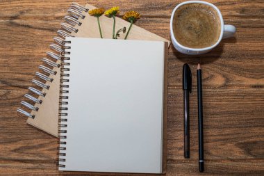 Üç çiçekli sarmal defterler. Bir tanesi beyaz çarşaf ve yanında siyah kalem ile açık. Tahta masada bir fincan daha kahve var..