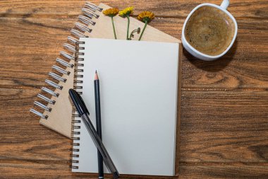 Üç çiçekli sarmal defterler. Üzerinde kalem ve kalem olan beyaz bir çarşaf. Tahta masada bir fincan daha kahve var..