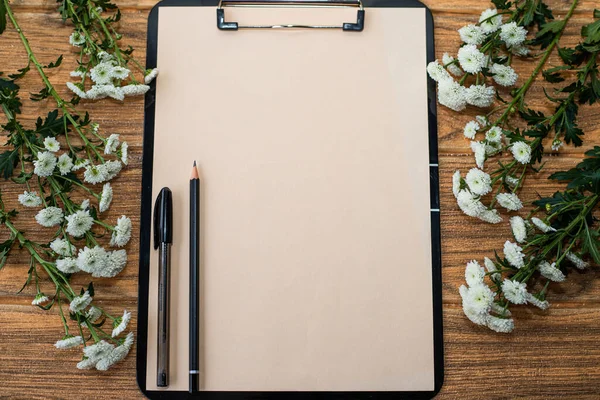 Black clipboard with ivory sheet, on it are a pen and a pencil. There are white flowers on both sides of the clipboard.