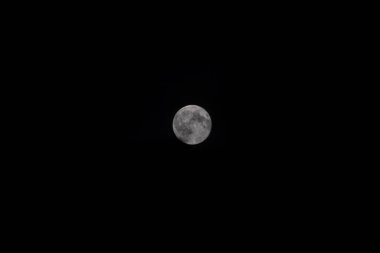 A close-up of the bright full Moon against the deep black night sky, with some clouds. The lunar disk is sharp and detailed, with the texture of the craters and lunar maria clearly visible.