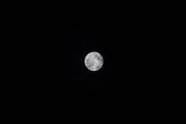 A close-up of the bright full Moon against the deep black night sky, with some clouds. The lunar disk is sharp and detailed, with the texture of the craters and lunar maria clearly visible.