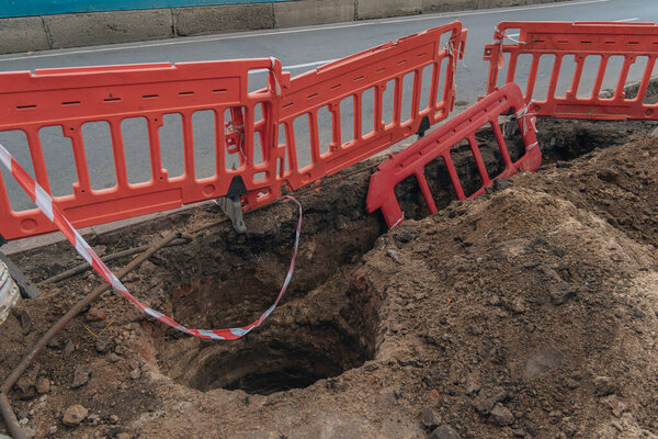 Крупный план глубокой, вырытой ямы (траншеи) на краю дороги, которая огорожена ярко-красными пластиковыми дорожными барьерами (секционное ограждение). Меры предосторожности при проведении дорожных работ