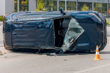 Devrilmiş mavi bir arabanın yakın çekimi. Kırık cam ve ezilmiş ceset görülebiliyor. Araba bir şehir caddesinin ortasında baş aşağı yatmaktadır. Kaza, sigorta talebi ve güvenlik uyumu.