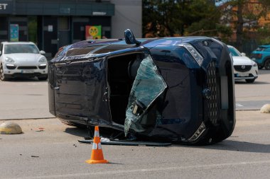 Devrilmiş mavi bir arabanın yakın çekimi. Kırık cam ve ezilmiş ceset görülebiliyor. Araba bir şehir caddesinin ortasında baş aşağı yatmaktadır. Kaza, sigorta talebi ve güvenlik uyumu.
