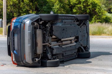 Devrilmiş mavi bir arabanın yakın çekimi. Kırık cam ve ezilmiş ceset görülebiliyor. Araba bir şehir caddesinin ortasında baş aşağı yatmaktadır. Kaza, sigorta talebi ve güvenlik uyumu.