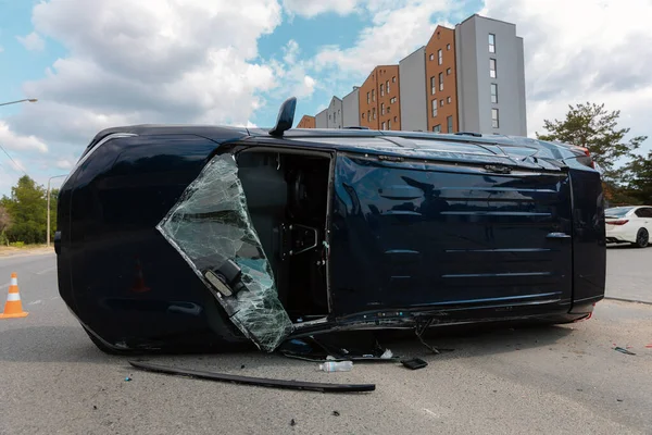 Devrilmiş mavi bir arabanın yakın çekimi. Kırık cam ve ezilmiş ceset görülebiliyor. Araba bir şehir caddesinin ortasında baş aşağı yatmaktadır. Kaza, sigorta talebi ve güvenlik uyumu.