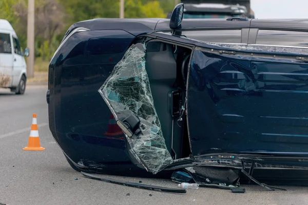 Devrilmiş mavi bir arabanın yakın çekimi. Kırık cam ve ezilmiş ceset görülebiliyor. Araba bir şehir caddesinin ortasında baş aşağı yatmaktadır. Kaza, sigorta talebi ve güvenlik uyumu.