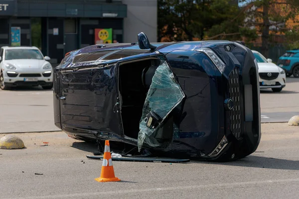Devrilmiş mavi bir arabanın yakın çekimi. Kırık cam ve ezilmiş ceset görülebiliyor. Araba bir şehir caddesinin ortasında baş aşağı yatmaktadır. Kaza, sigorta talebi ve güvenlik uyumu.