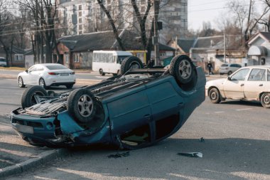 Bir trafik kazasından sonra mavi bir yolcu arabası kaldırımlı bir yolda baş aşağı yatıyor. Şasinin yakın çekimi gösteriliyor. Güvenlik, sigorta, trafik ve sonuçlar konuları.
