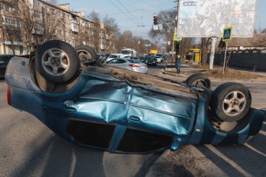 Bir trafik kazasından sonra mavi bir yolcu arabası kaldırımlı bir yolda baş aşağı yatıyor. Şasinin yakın çekimi gösteriliyor. Güvenlik, sigorta, trafik ve sonuçlar konuları.